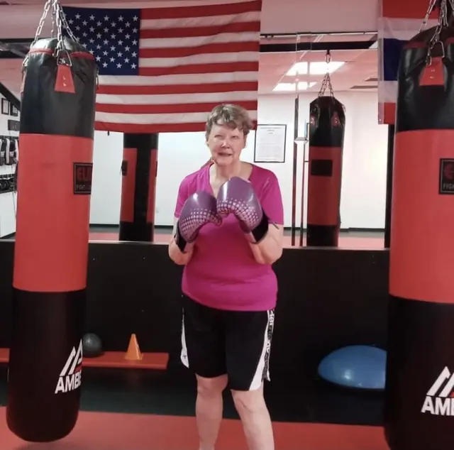 A smiling student in pink gloves between the heavy bags